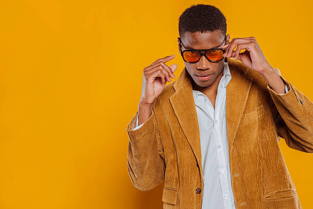 Confident businessman adjusting orange sunglasses while wearing brown corduroy jacket and white shirt against a vibrant yellow background.