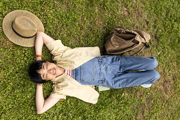 High angle shot of smiling Asian woman lying on grass with eyes closed while relaxing in nature