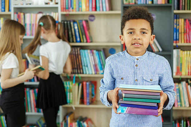 Young boy stands in a vibrant school library holding a colorful stack of books, while other children browse the shelves behind.