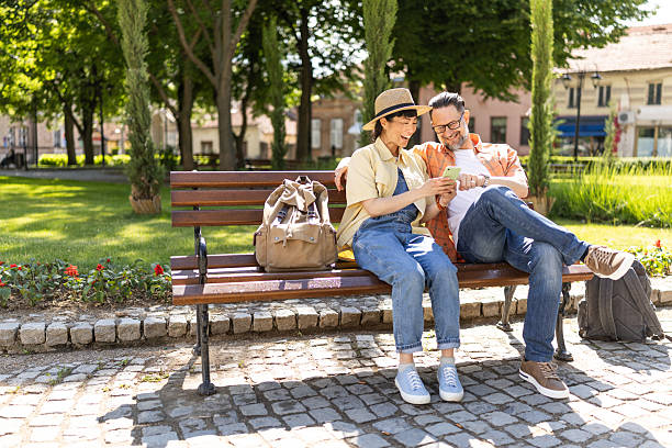 Kazan, Russia - June 9, 2023: Summer afternoon in city public park. Senior asian woman sitting on bench in shade and using mobile phone. Lifestyle, technology concept