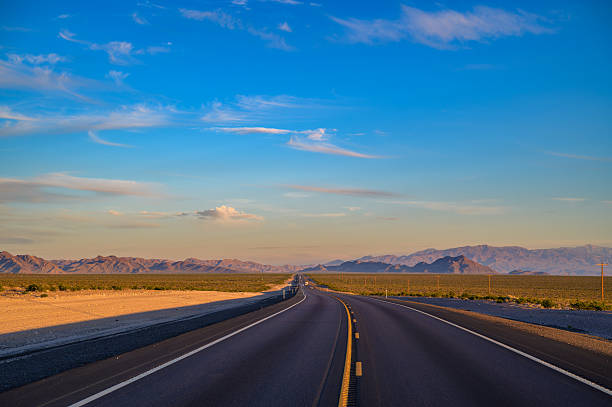 A two-lane road stretches into the distance toward Death Valley National Park, framed by desert terrain and distant mountains under a colorful sunset sky.