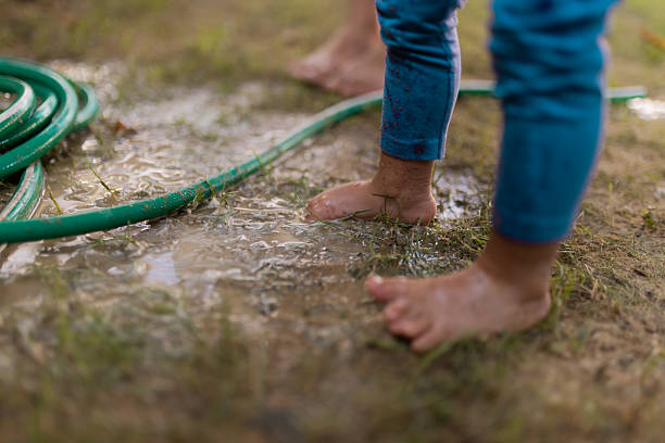 Barefoot children enjoy playing in wet, muddy puddles alongside a green garden hose