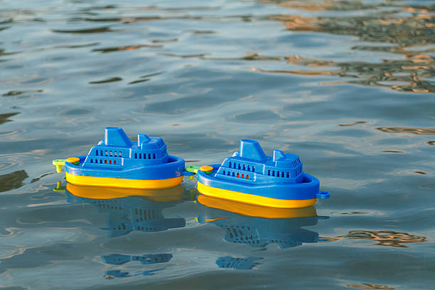 Two blue and yellow plastic toy boats float on a calm water surface. Gentle ripples reflect the sunlight, creating a peaceful and playful scene.