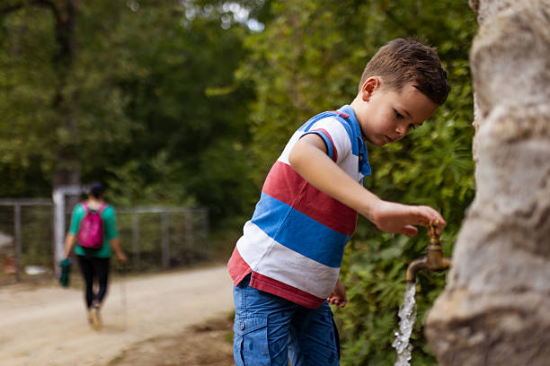 A child in casual attire operates an outdoor water faucet while in a natural park setting, with trails and foliage in the background, creating a scene of refreshment and outdoor activity.