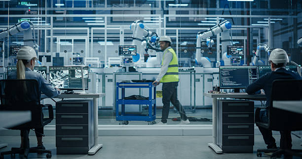 istock Establishing Shot at an Electronics Factory with Robotic Arms. Diverse Engineers and Developers Working on Desktop Computers. Indian Worker in High Visibility Vest Pushing a Trolley with Equipment 2217640925