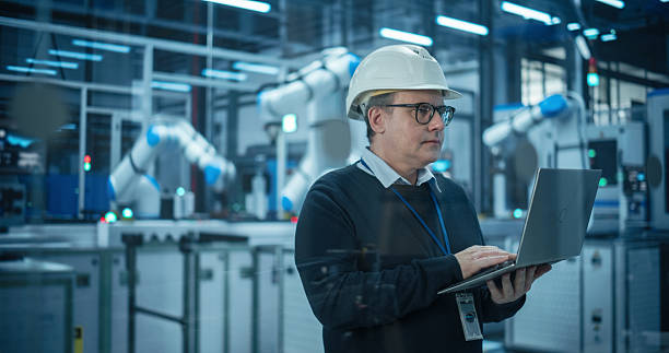 istock Adult Caucasian Engineer in a Hard Hat Working in a Modern Industrial Facility, Using Laptop Computer to Monitor Robotic Arms in a Futuristic Electronics Manufacturing Factory 2217640971