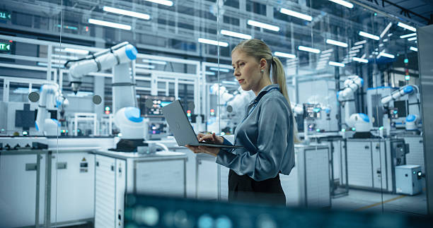 istock Caucasian Female Engineer Using a Laptop Computer to Monitor Robotic Arms in an Advanced Industrial Factory. Specialist Overlooking Modern Automated Electronics Manufacturing 2217641581