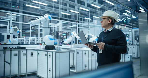 istock Industrial Systems Administrator Working on Laptop Computer at a Modern Manufacturing Plant. Software Developer Programming the Work of the Robotic Arms, Monitoring Production on the Assembly Conveyor 2217641712