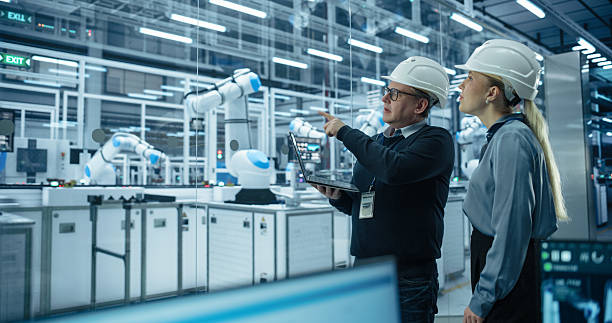 istock Male Engineer and Female Project Manager Standing Next to an Assembly Line, Talking, Using Laptop and Monitoring Production at a Modern Automated Electronics Manufacture with Robotic Arms 2217641875