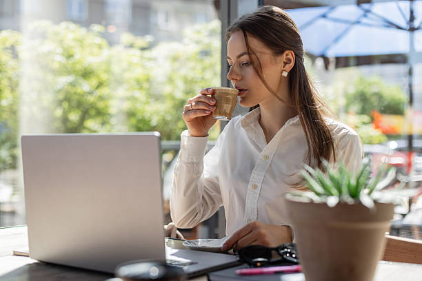 istock Young businesswoman drinking coffee while working on a laptop at a bright modern cafe. Remote work, freelance lifestyle, productivity and relaxation concept. 2217644209