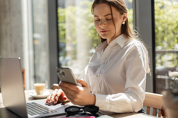 istock Confident female employee handling digital tasks on phone and computer in a contemporary office setting. Office lifestyle: woman checking phone while working at laptop. Bright interior with natural li 2217644290
