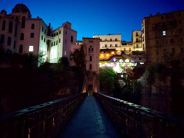 Night view of the Mellah Slimane Bridge in Constantine, Algeria. The suspension bridge, softly illuminated, leads towards a cluster of historic buildings perched on the cliffside.