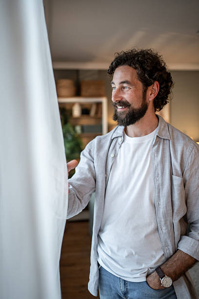 istock Smiling bearded man opening curtains and looking out window at home 2217650241