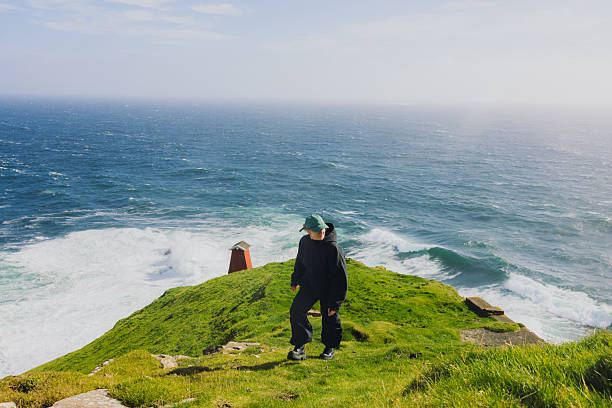 Front view of active female explorer hiking at the edge of the cliff overlooking majestic ocean during sunny day on Faroe Islands