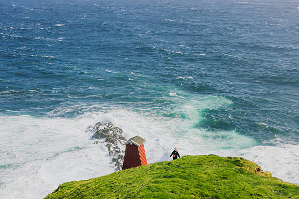 Rear view of female explorer in distant staying at the edge of the cliff overlooking majestic ocean during sunny day on Faroe Islands