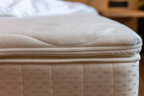 istock A padded mattress is positioned on a wooden bed frame, featuring a smooth, soft top layer. The cozy bedroom is adorned with warm lighting and inviting linens, creating a relaxing atmosphere 2217677747