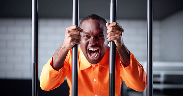 Young Man In Orange Prison Uniform Locked Behind Jail Bars. Accused Criminal Serving Term In Isolation Cell
