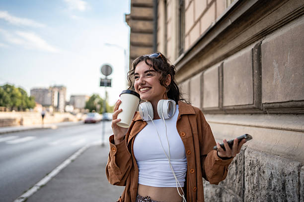 A happy young woman wearing headphones, enjoying coffee, and using her smartphone while outdoors in an urban environment.