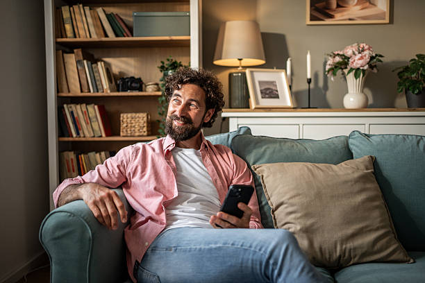 istock Smiling man relaxing on sofa holding smartphone and looking away 2217697583
