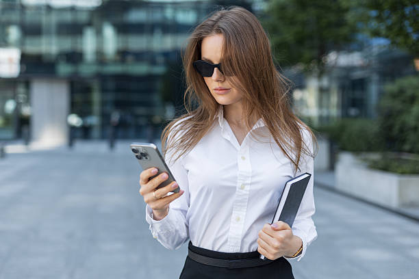 istock Confident young businesswoman in sunglasses using smartphone while holding a notebook. Professional urban setting with modern glass buildings in the background 2217707928