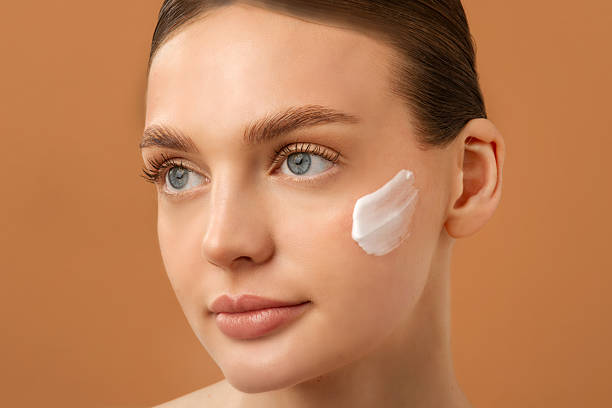 istock Young woman applying moisturizer on cheek, using under eye treatment, posing over beige background. Pretty lady with swatch of cream on cheek 2217719370