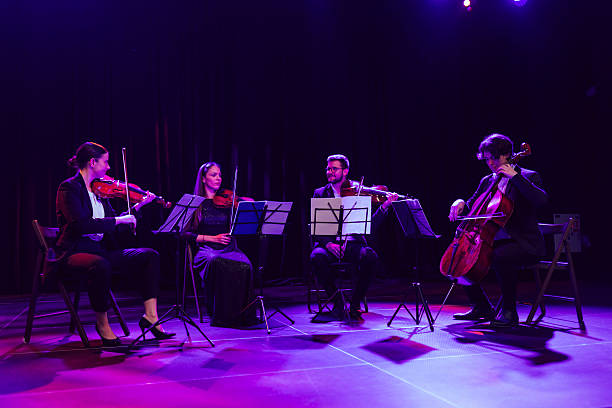 istock Four string quartet musicians play viola, cello and violins while sitting next to each other and performing classical music on a theater stage 2217726385