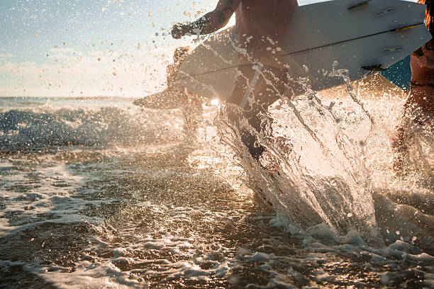 Caucasian male surfers enthusiastically dash into the sunrise-lit waves of Gold Coast beach, carrying surfboards. Wearing swimwear, they embody the thrill of the ocean adventure, blending passion and sport in this breathtaking coastal location.