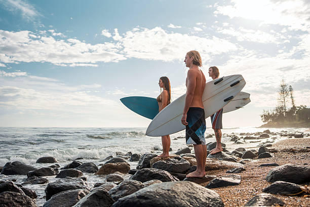 Caucasian male and female surfers eagerly observe ocean waves at sunrise. Dressed in swimwear and holding surfboards, they stand on rocky terrain by the coast, ready for the surf in this picturesque seaside location.