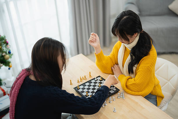 Two Women Playing Chess at Home During Daytime with Natural Light Streaming Through the Window, Cozy Atmosphere and Focused Expressions