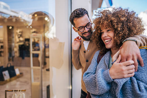 Young couple hugging and smiling while admiring a shop window, holding colorful shopping bags and enjoying a delightful day out together in the vibrant city atmosphere