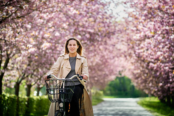 A twenty-year-old confident young woman riding her retro-style bike through an avenue of blooming cherry blossom trees. She is wearing a fashionable Burberry-style trench coat, paired with a grey sweater, black trousers, and has a pale complexion, complemented by long, chestnut-coloured hair.