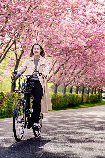 A twenty-year-old confident young woman riding her retro-style bike through an avenue of blooming cherry blossom trees. She is wearing a fashionable Burberry-style trench coat, paired with a grey sweater and black trousers. She has a pale complexion, complemented by long, chestnut-coloured hair.