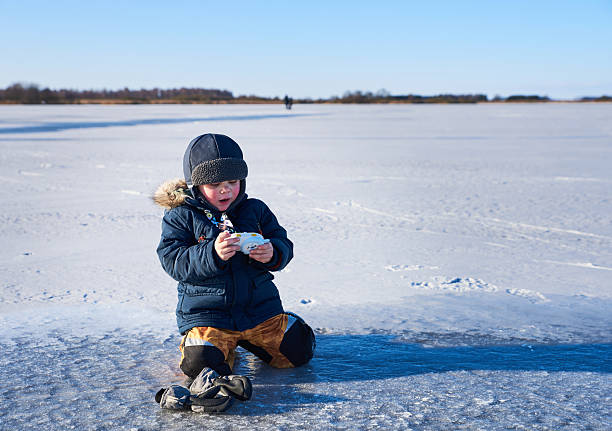 A young child explores curiosity and play on a frozen lake, kneeling in snow while using a toy camera. The clear sky and vast icy landscape highlight themes of wonder, discovery, and solitude