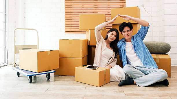 istock Happy young couple sitting with moving boxes, forming a roof shape with hands, symbolizing a fresh start in a new home. Perfect image for relocation, real estate, or home moving service concepts. 2217781767