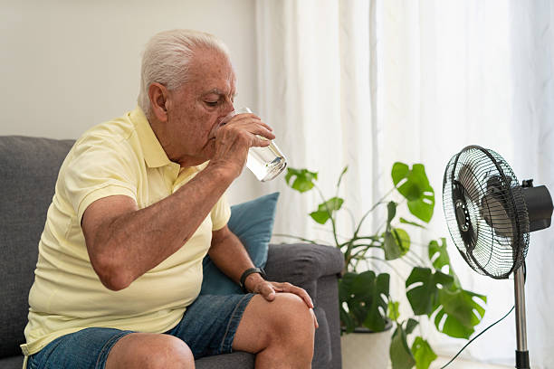 Senior man refreshing himself drinking water and using an electric fan during a hot summer at home