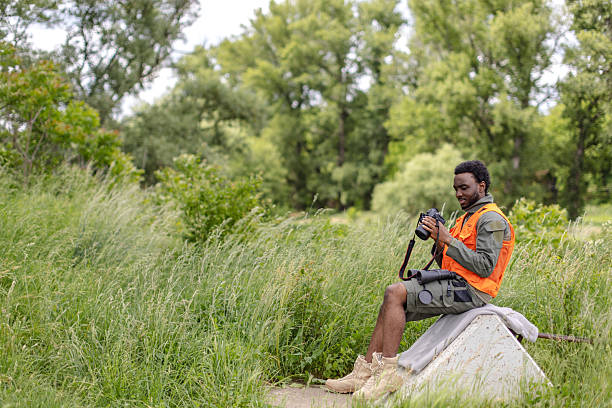 A young man wearing an orange vest sits comfortably outdoors, observing wildlife on his camera in a beautiful forest. His cheerful demeanor reflects joy in birding and exploration amidst nature.