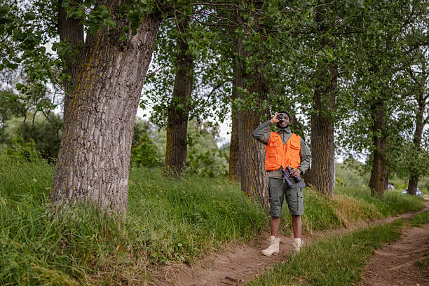 In a vibrant forest setting, a young man dressed in orange is engaged in bird watching. He stands near trees, holding binoculars, filled with curiosity and joy for nature.