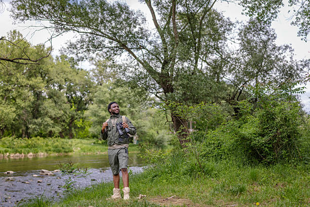 A young man stands in a lush green forest, exploring nature with binoculars in hand. Surrounded by trees and a nearby river, he smiles in the joy of bird watching and outdoor exploration.