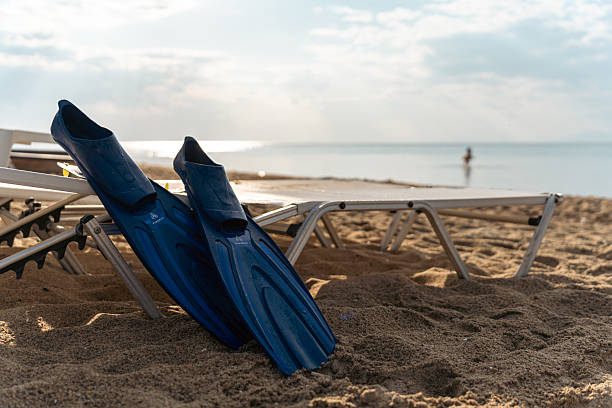 A pair of blue swimming fins on the beach sand, alongside chairs, with a serene backdrop of the sea and bright sky.
