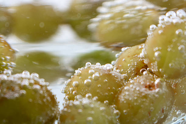 Green grapes submerged in a sparkling liquid, covered with numerous tiny bubbles. The grapes appear fresh, with light green skins and slight translucence. The background is a bright, plain white, enhancing the contrast and highlighting the effervescence. The image captures the refreshing essence of the fruit, suggesting freshness and vibrancy, often associated with healthy beverages or recipes.