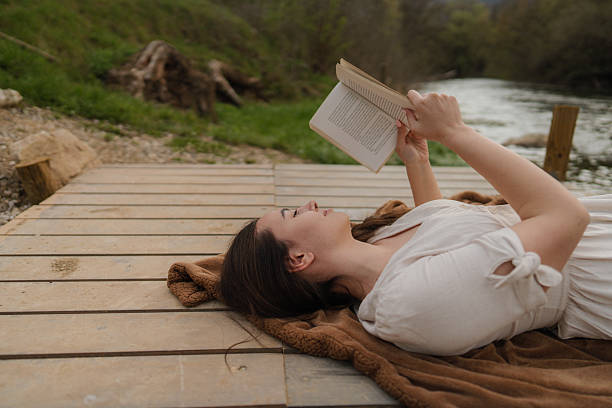 Photo of a young woman reading, soaking the sun and relaxing on a riverside dock