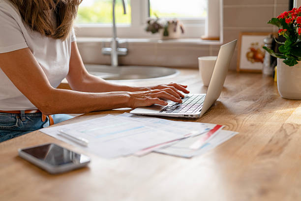 Home office workspace with a woman typing on a laptop amidst paperwork and a smartphone while enjoying a bright morning