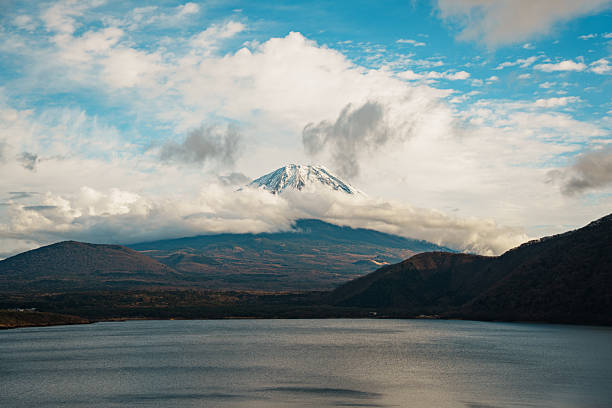 Mount Fuji majestically rises behind Lake Motosuko, its snow capped peak framed by rolling hills and dramatic clouds under a vibrant blue sky.