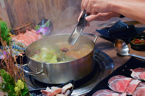 istock Person's hands using tongs to add ingredients into a steaming hot pot with various fresh meats, seafood, and vegetables arranged on a wooden table. 2217831085