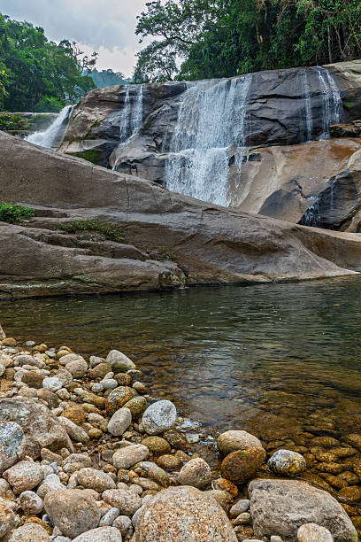 istock A multi-tiered waterfall cascades over large granite rocks into a clear natural pool, surrounded by lush green tropical forest under a cloudy sky. 2217831484