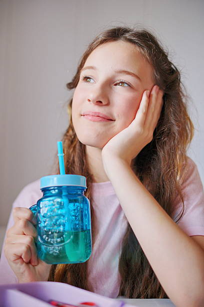 A girl with long hair smiles gently while holding a blue cup, lost in thought during a relaxing afternoon at home.