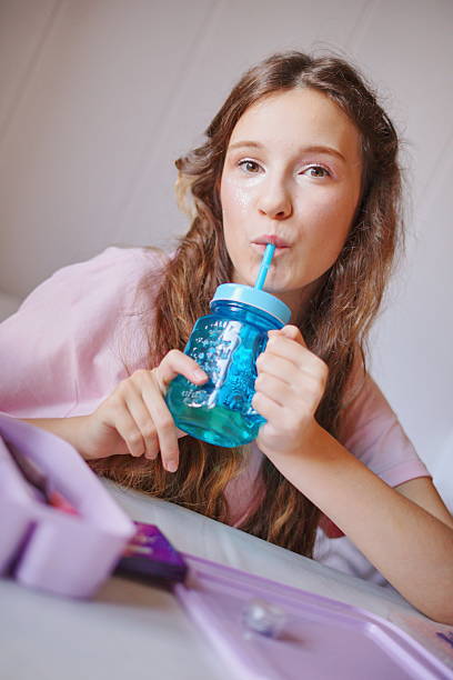 Cute girl enjoying a refreshing drink while sitting on her bed with school supplies around her during the afternoon