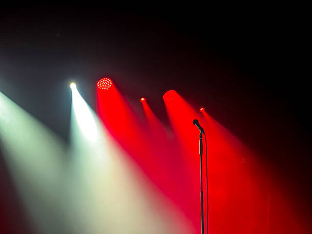 istock Microphone on stand receives intense red and white spotlight beams on dark stage, representing live shows, music concerts, public speaking, and dramatic performance atmosphere 2217846726