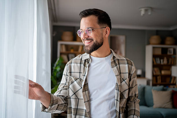 istock Smiling young man opening curtains at home and looking out 2217849957