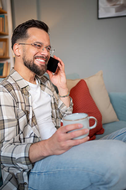 istock Smiling man talking on phone and drinking coffee on sofa 2217850828
